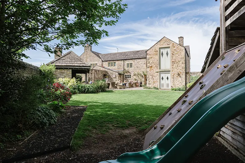 Relaxing garden area at The Fleece Inn with stone buildings, lawn and children’s play slide